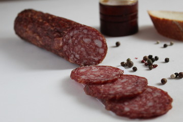 a stick of smoked raw sausage and slices cut into rings on a white background with peppers peas and baguette and a salt shaker in the background