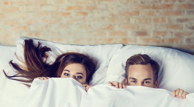 Young playfull attractive couple peeping from bedsheet on the bed at bedroom. Love, relationship, dating, happy people, bedtime concept picture.