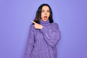 Young beautiful woman wearing casual turtleneck sweater standing over purple background Surprised pointing with finger to the side, open mouth amazed expression.