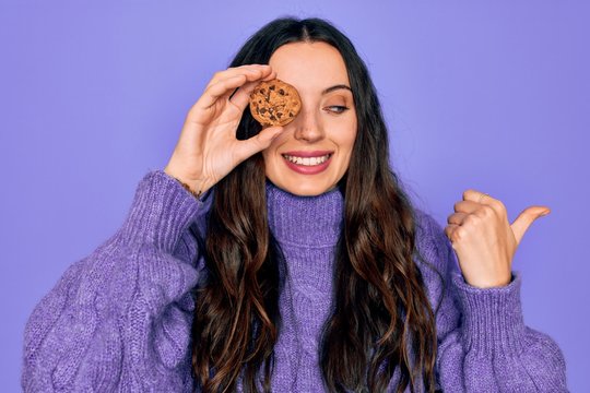 Young beautiful woman with blue eyes holding chocolate sweet coockie over eye pointing and showing with thumb up to the side with happy face smiling