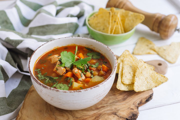 Chicken soup on chicken broth, with nachos, chickpeas and tomatoes in a deep beige bowl. Sprinkled with chopped parsley leaves. On a wooden table, a cutting board, with a pepper mill, a kitchen towel.
