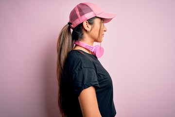 Fototapeta premium Young beautiful fitness sports woman wearing training cap and headphones over pink background looking to side, relax profile pose with natural face with confident smile.
