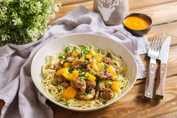 Deep plate with spaghetti, minced meat, chopped parsley, yellow bell pepper. Nearby is a bowl with sauce, a fork and a knife, a table napkin, a kitchen waffle towel. Serving per person.