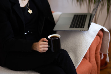 Fashionable  woman sitting on sofa at home and working from home, remotely. She is keyboarding on laptop and holding  coffee. Freelance work concept. Copy space for text. Close up of laptop and hands.