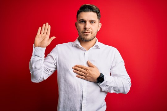 Young business man with blue eyes wearing elegant shirt standing over red isolated background Swearing with hand on chest and open palm, making a loyalty promise oath