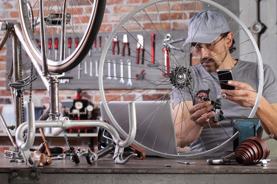 man repair the vintage bicycle with mobile phone at the computer search spare parts on the internet in garage workshop on the workbench with tools, diy concept