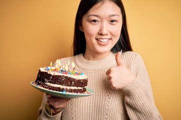 Young asian woman holding birthday cake with candles burning over yellow isolated background happy with big smile doing ok sign, thumb up with fingers, excellent sign