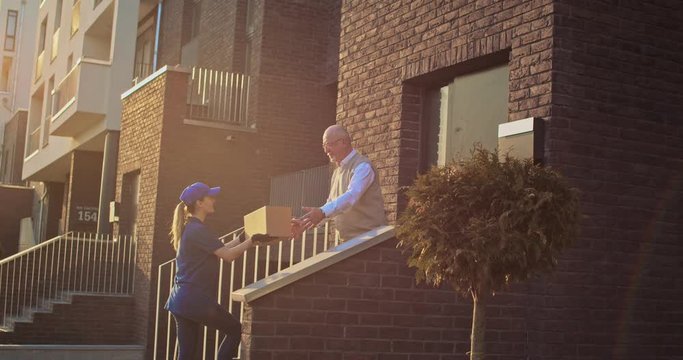 Young Caucasian girl bringing carton box to old male pensioner and handing outdoors. Senior man getting parcel at his house outdoor from female courier.