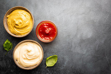 Classic set of sauces, American yellow mustard, ketchup, barbecue sauce, mayonnaise on black stone background, top view with copy space.