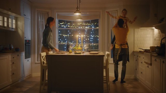 Happy Family of Three Cooking and Having Dinner Together. Mother Prepares Food, Cute Little Girl Runs to Father, Hugs Him, They Dance and Swirl. Festive Table in Cozy Kitchen