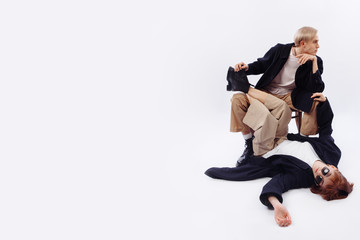 Classic style in clothing. The blond guy is sitting on a chair. He's wearing a black coat, a white t-shirt, and beige straight pants. The girl with glasses is lying in the guy's legs. 