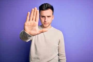 Fototapeta premium Young handsome caucasian man wearing casual sweater over purple isolated background doing stop sing with palm of the hand. Warning expression with negative and serious gesture on the face.