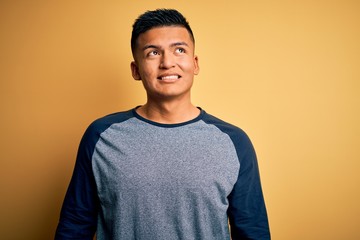 Young handsome latin man wearing casual t-shirt standing over yellow background looking away to side with smile on face, natural expression. Laughing confident.