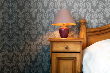 Close up of a wooden bed with white sheets and a cozy lamp, pattern wallpaper retro design.