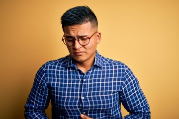 Young handsome latin man wearing casual shirt and glasses over yellow background with hand on stomach because indigestion, painful illness feeling unwell. Ache concept.