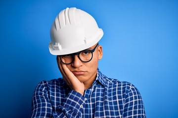 Young handsome engineer latin man wearing safety helmet over isolated blue background thinking looking tired and bored with depression problems with crossed arms.