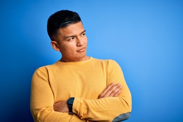 Young handsome latin man wearing yellow casual sweater over isolated blue background looking to the side with arms crossed convinced and confident