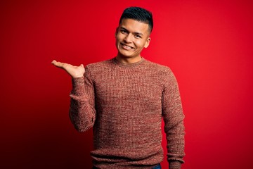 Young handsome latin man wearing casual sweater standing over red background smiling cheerful presenting and pointing with palm of hand looking at the camera.