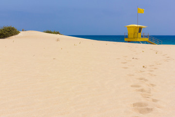 Fototapeta na wymiar Footprints on sandy beach leading towards yellow lifeguard station on sunny day in Fuerteventura island. Sand dune by baywatch tower at Corralejo natural park. Summer travel destination concept