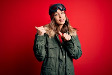 Young blonde girl wearing ski glasses and winter coat for ski weather over red background Pointing to the back behind with hand and thumbs up, smiling confident