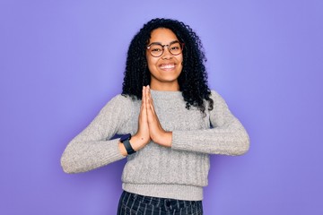 Young african american woman wearing casual sweater and glasses over purple background praying with hands together asking for forgiveness smiling confident.