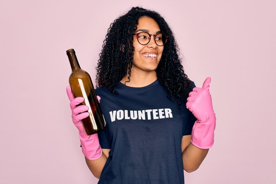African american curly woman wearing volunteer t-shirt doing volunteering recycling glass bottle pointing and showing with thumb up to the side with happy face smiling