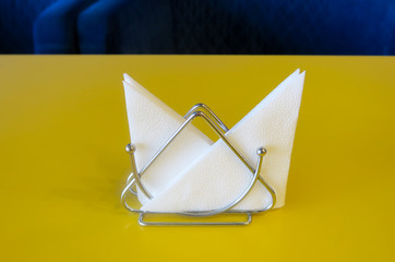 White napkins in a napkin holder stand on a yellow table. Dark blue background.