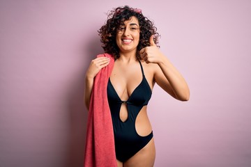 Young beautiful curly arab woman on vacation wearing swimsuit holding beach towel happy with big smile doing ok sign, thumb up with fingers, excellent sign