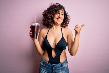 Beautiful arab woman on vacation wearing swimsuit drinking cola refreshment using straw pointing and showing with thumb up to the side with happy face smiling
