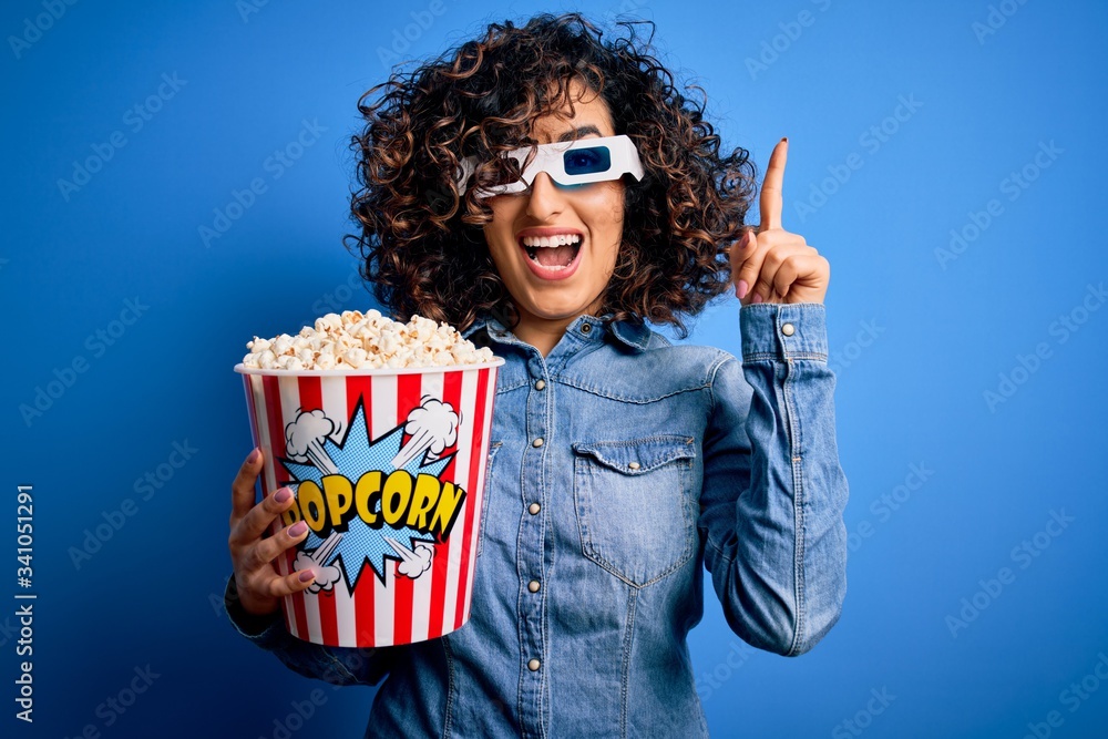 Sticker Young beautiful curly arab woman watching movie using 3d glasses eating popcorn snack surprised with an idea or question pointing finger with happy face, number one