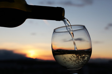 White wine pouring from a bottle into the glass on beautiful sunset background, orange sun is shining through the dark clouds. Concept of celebration, romantic dinner