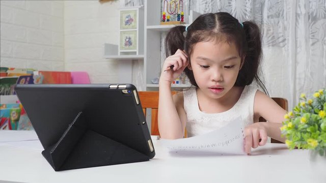 Asian little cute girl 6 years old studying online with tablet and writing in paper on white table. Preschool lovely kid learning online at home.