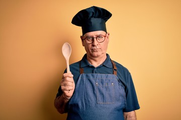 Middle age cooker man wearing apron and hat holding wooden spoon over yellow background with a...