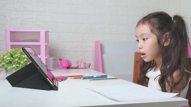 Asian little cute girl 6 years old studying online with tablet and learning pronunciation of words with teacher on white table. Preschool lovely kid learning online at home.