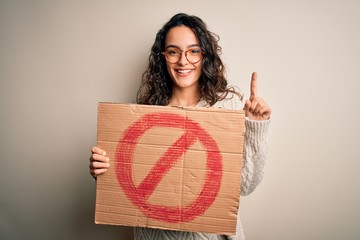 Young beautiful woman with curly hair holding banner with prohibited signal message surprised with an idea or question pointing finger with happy face, number one