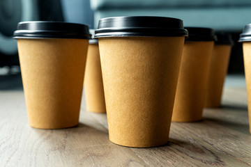 A line of disposable coffee cups on a wooden table. Brown paper cup with black lid 