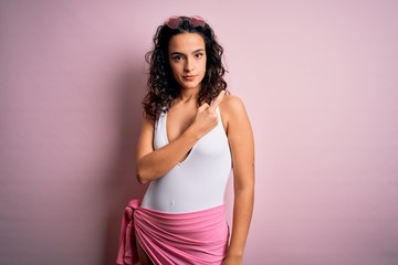 Beautiful woman with curly hair on vacation wearing white swimsuit over pink background Pointing with hand finger to the side showing advertisement, serious and calm face