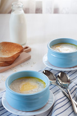 Oatmeal milk porridge with butter in blue bowls on a textile napkin, bread toast and a bottle of milk on a white table against the background of a window with curtains