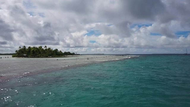 A spectacular low tracking shot of the blue turquoise lagoon and coral reef break on a cloudy day with small islands filled with palm treesd blow in the wind aerial drone shot in Rangiroa, French Poly