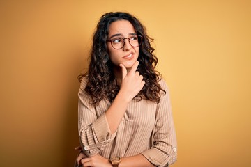 Beautiful woman with curly hair wearing striped shirt and glasses over yellow background Thinking worried about a question, concerned and nervous with hand on chin