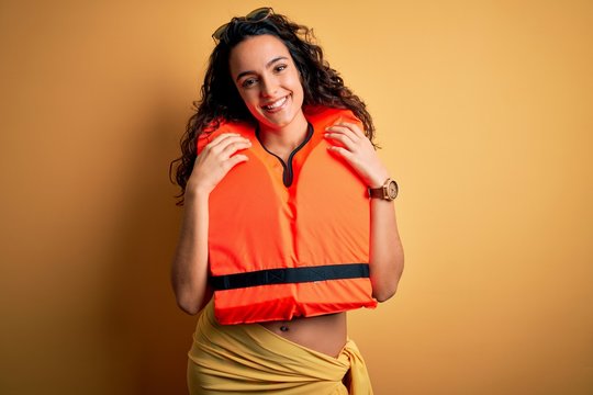 Young beautiful woman with curly hair wearing orange lifejacket over yellow background with a happy and cool smile on face. Lucky person.