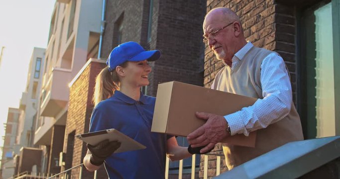 Caucasian girl bringing carton box to male pensioner standing outdoor at house. Delivering service concept. Female courier deliver parcel to old man in glasses and he meeting her outside house..