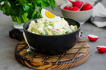 Potato salad with radish, eggs, green peas, sour cream and mayonnaise in a bowl on a board on a dark concrete background. Horizontal orientation, close up.