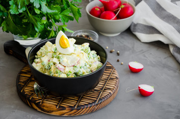 Potato salad with radish, eggs, green peas, sour cream and mayonnaise in a bowl on a board on a dark concrete background. Horizontal orientation, copy space.