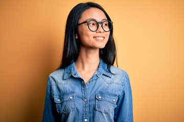 Young beautiful chinese woman wearing casual denim shirt over isolated yellow background looking away to side with smile on face, natural expression. Laughing confident.