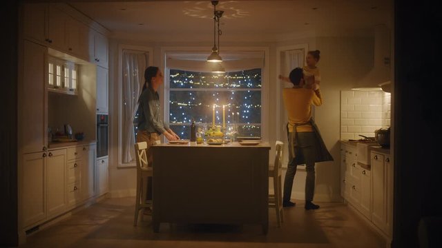 Family of Three Cooking and Having Dinner Together. Mother Prepares Food, Little Girl Runs to Father, Hugs Him and They Dance. Festive Table in Stylish Kitchen Interior with Warm Light. Slow motion 