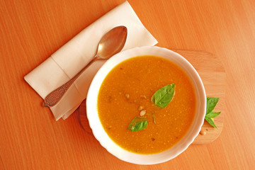 Top view of a plate of pumpkin soup with basil leaves and pine nuts on wooden plate and green background with a spoon