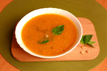 Top view of bowl of pumpkin soub with basil leaves and pine nuts on wooden plate and green background