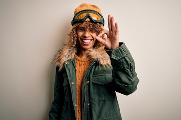 Young african american skier woman with curly hair wearing snow sportswear and ski goggles smiling positive doing ok sign with hand and fingers. Successful expression.