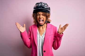 African american motorcyclist woman with curly hair wearing moto helmet over pink background celebrating mad and crazy for success with arms raised and closed eyes screaming excited. Winner concept